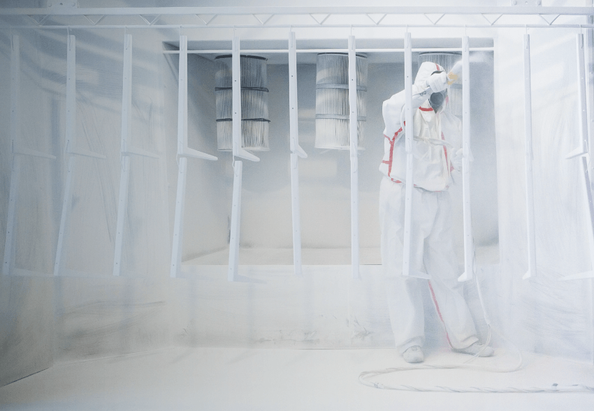 A man wearing personal protective equipment while powder coating eight metal wall brackets and two large air filters in the background.