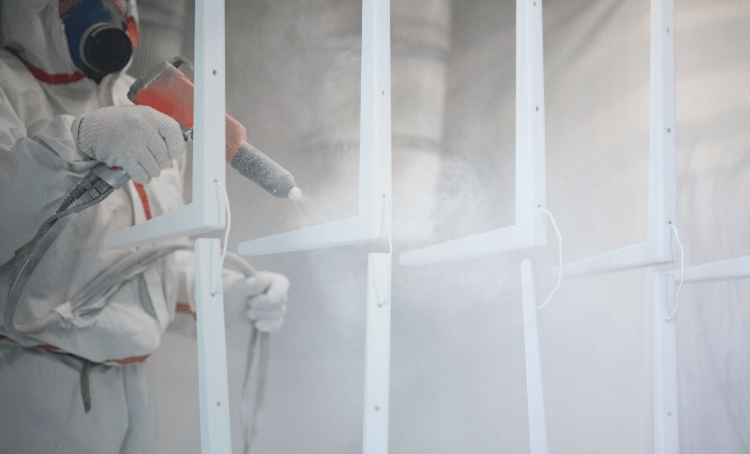 A man wearing personal protective equipment while powder coating eight metal wall brackets.