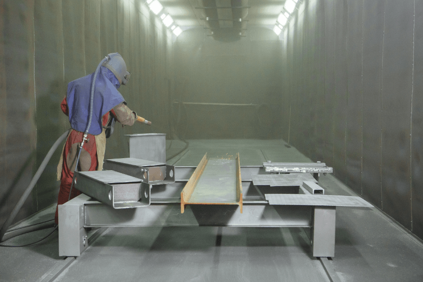 A man wearing personal protective equipment while shot blasting four metal beam to remove contaminants. 